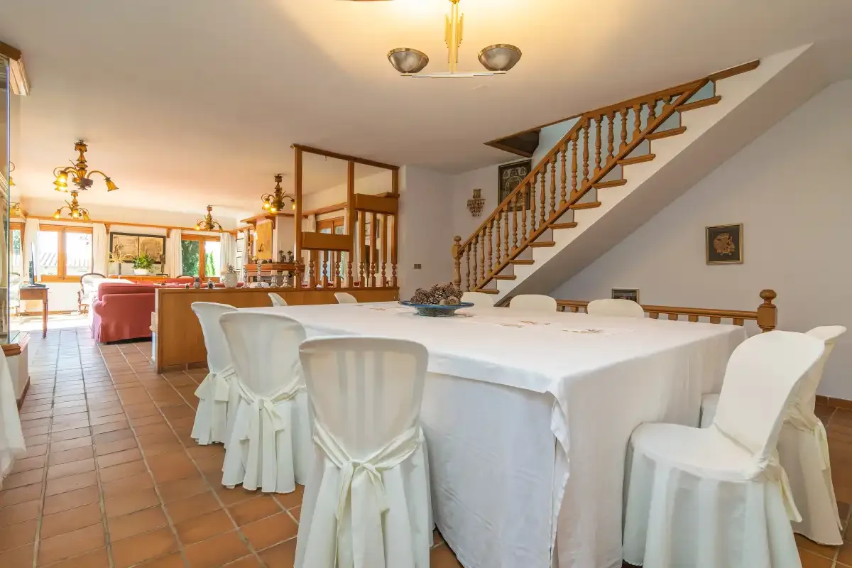 A dining area with a long table and covered chairs beneath a staircase.