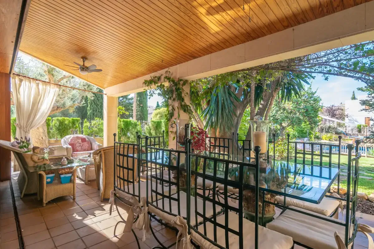 A covered patio with seating overlooks a garden and swimming pool.