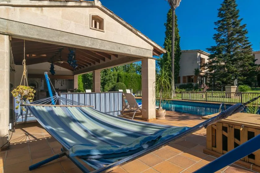 A covered patio beside a swimming pool, with a hammock in the foreground.