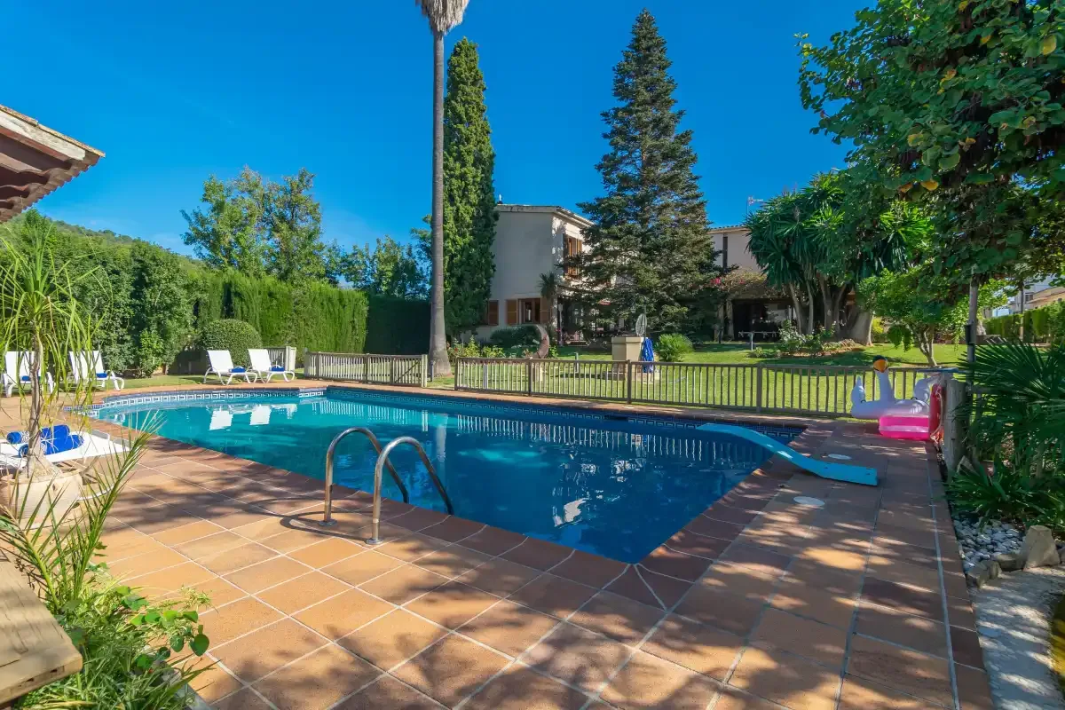 An outdoor swimming pool is surrounded by a tiled patio, loungers, and greenery.
