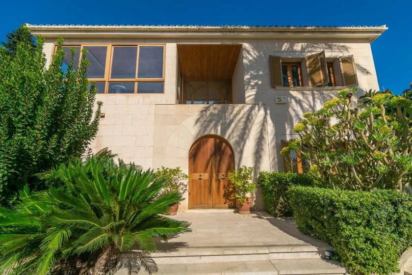 A pale stone house façade with an arched wooden entrance and garden plants.
