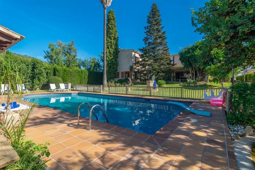 An outdoor swimming pool is surrounded by a tiled deck, lounge seating, and greenery.