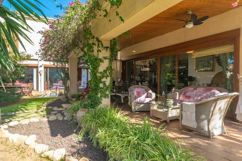 A covered outdoor seating area beside a garden, with cushioned chairs and a table.