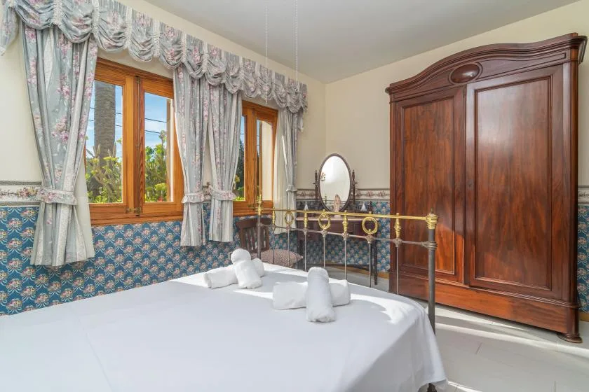 A bedroom with a neatly made bed beside curtained windows and a wooden wardrobe.