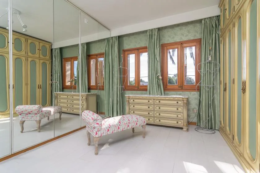 A bedroom with patterned seating, wooden dressers, and windows framed by green curtains.