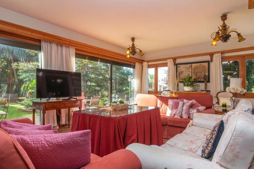 A bright living room with sofas, a television, and large windows overlooking greenery.