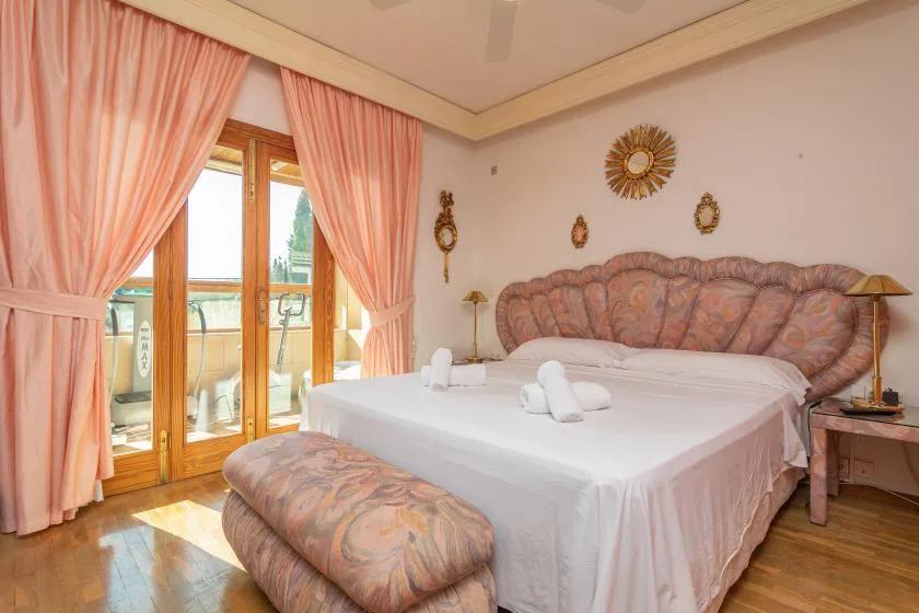 A bedroom with a large bed, patterned headboard, and pink curtains by the balcony doors.