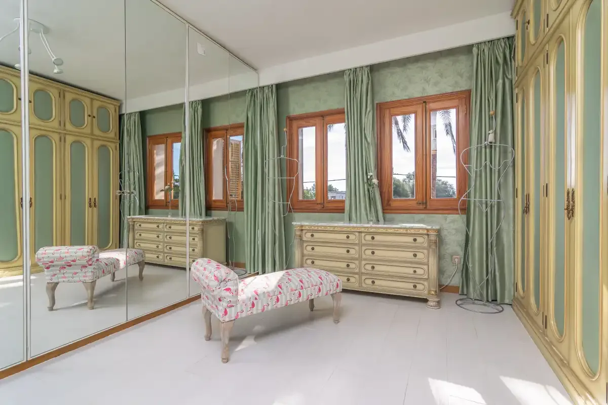 A bedroom with mirrored wardrobes, floral seating, and windows framed by green curtains.