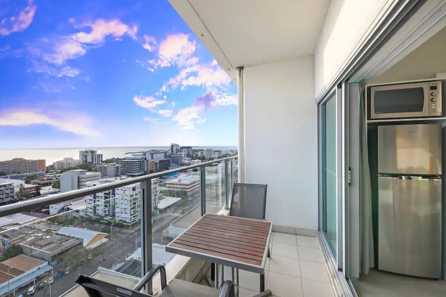A balcony with an outdoor table overlooks city buildings and the distant coastline.