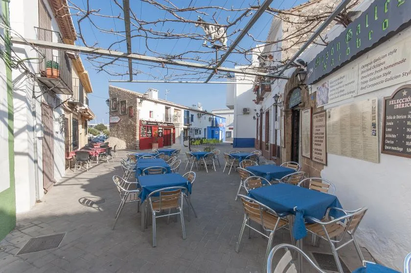 Una zona de comedor al aire libre tiene mesas con manteles azules y asientos bajo una pérgola metálica.