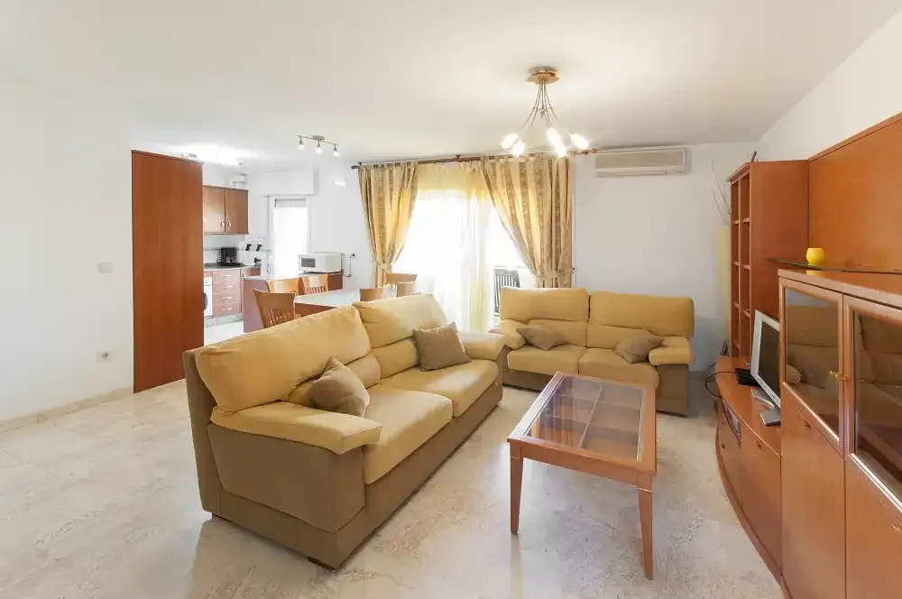 Una sala de estar con asientos beige y una mesa de centro con tapa de vidrio.