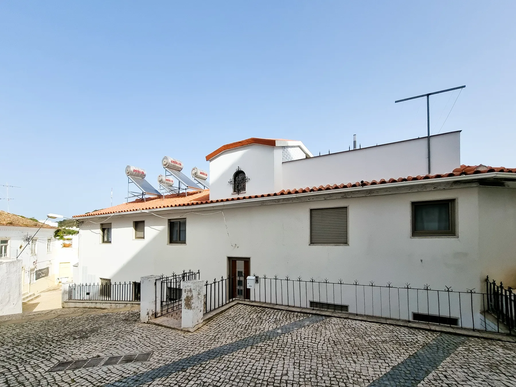 A white low-rise building with a tiled roof borders a cobblestone courtyard.