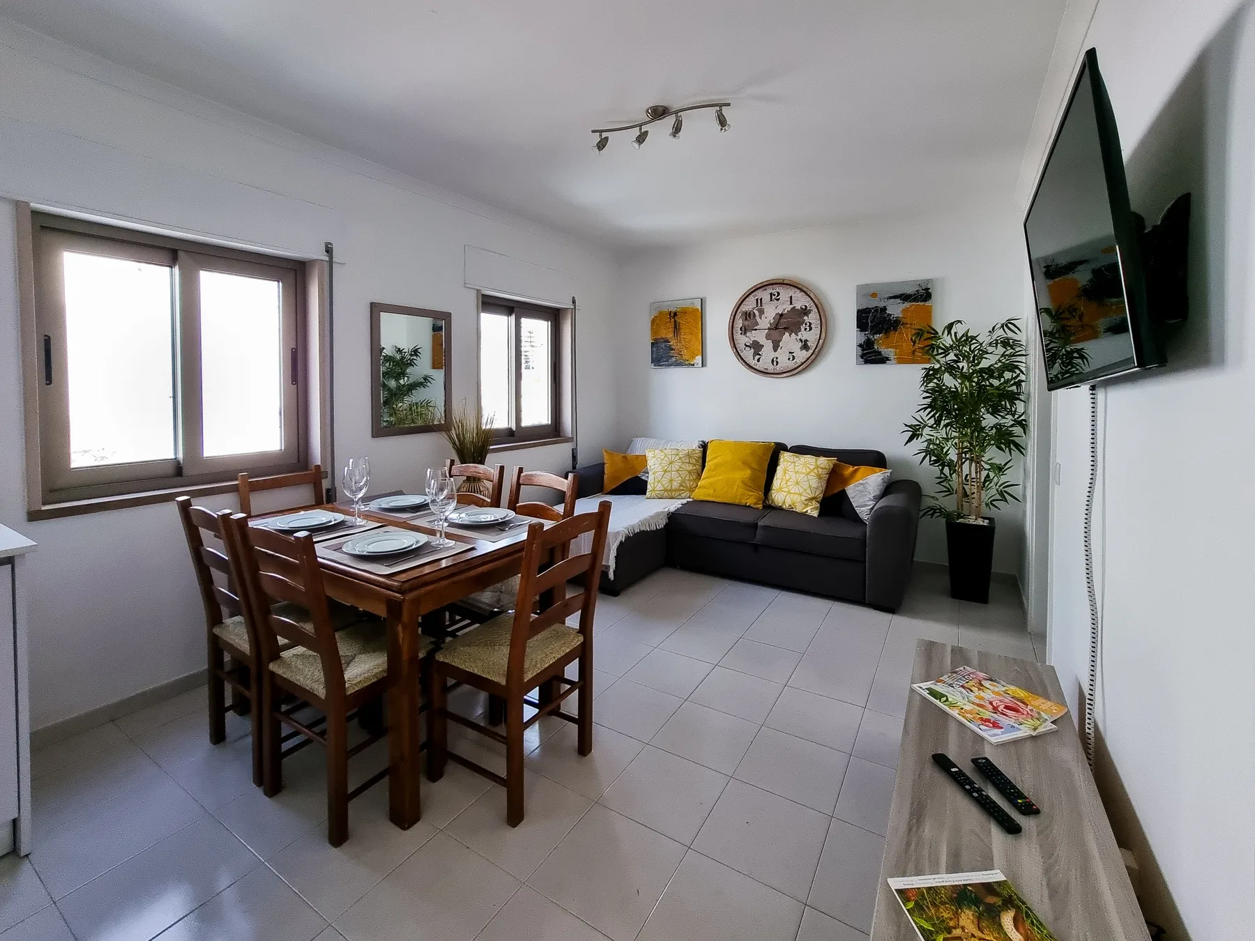 Sala de estar y comedor con un sofá oscuro, una mesa de comedor y un televisor en la pared.
