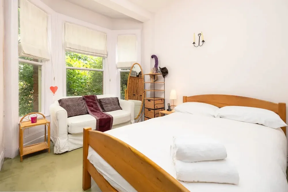 A bright bedroom with a white bed and a seating area beside tall windows.