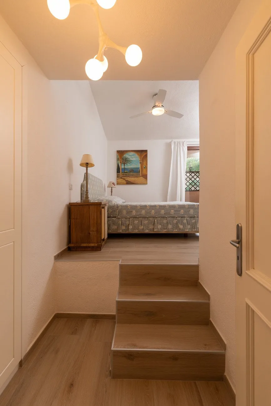 A bedroom is visible beyond a short set of wooden steps, with a bed and ceiling fan.
