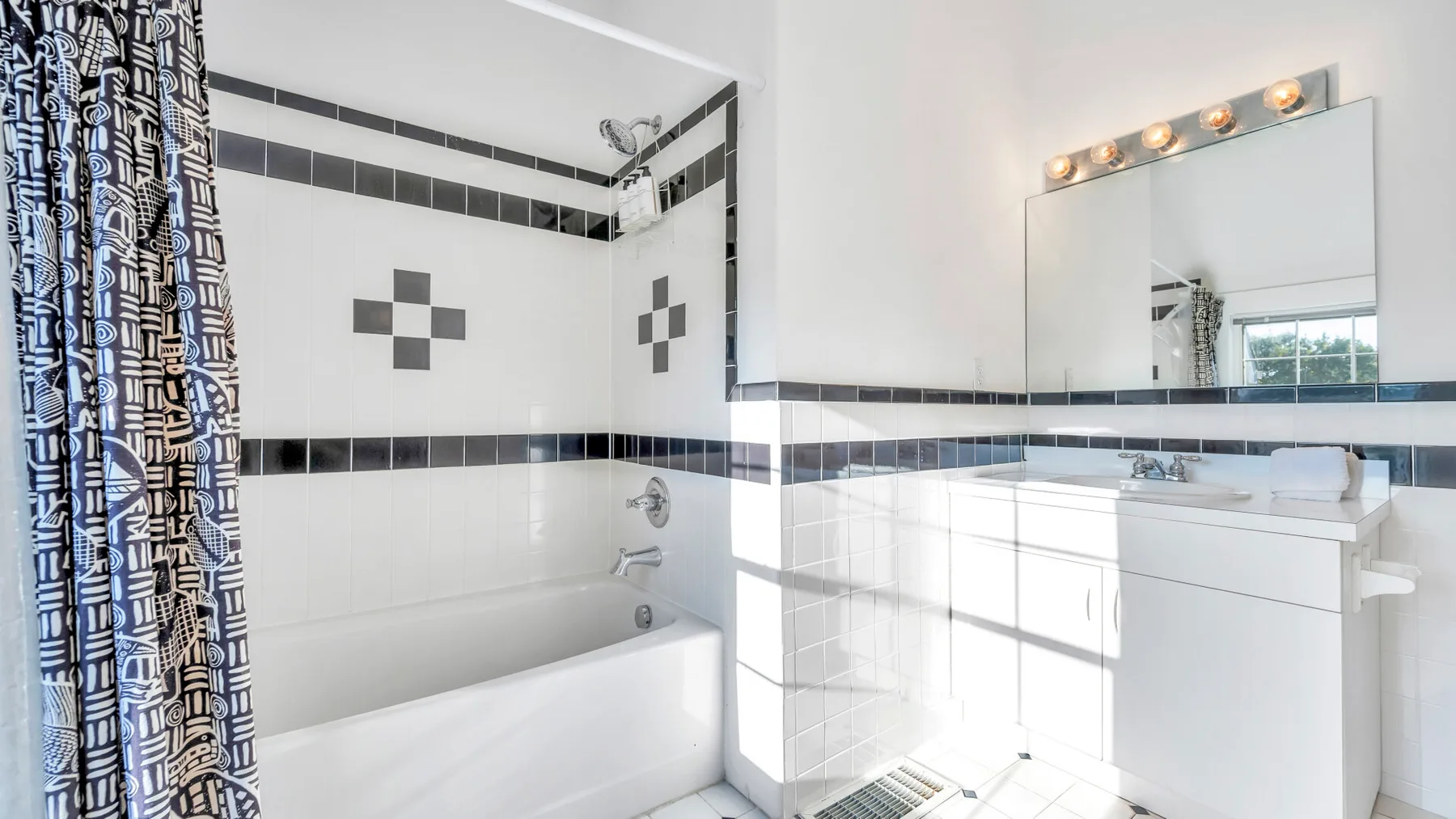 Un baño luminoso con bañera, cortina de ducha estampada y lavabo bajo un espejo grande.