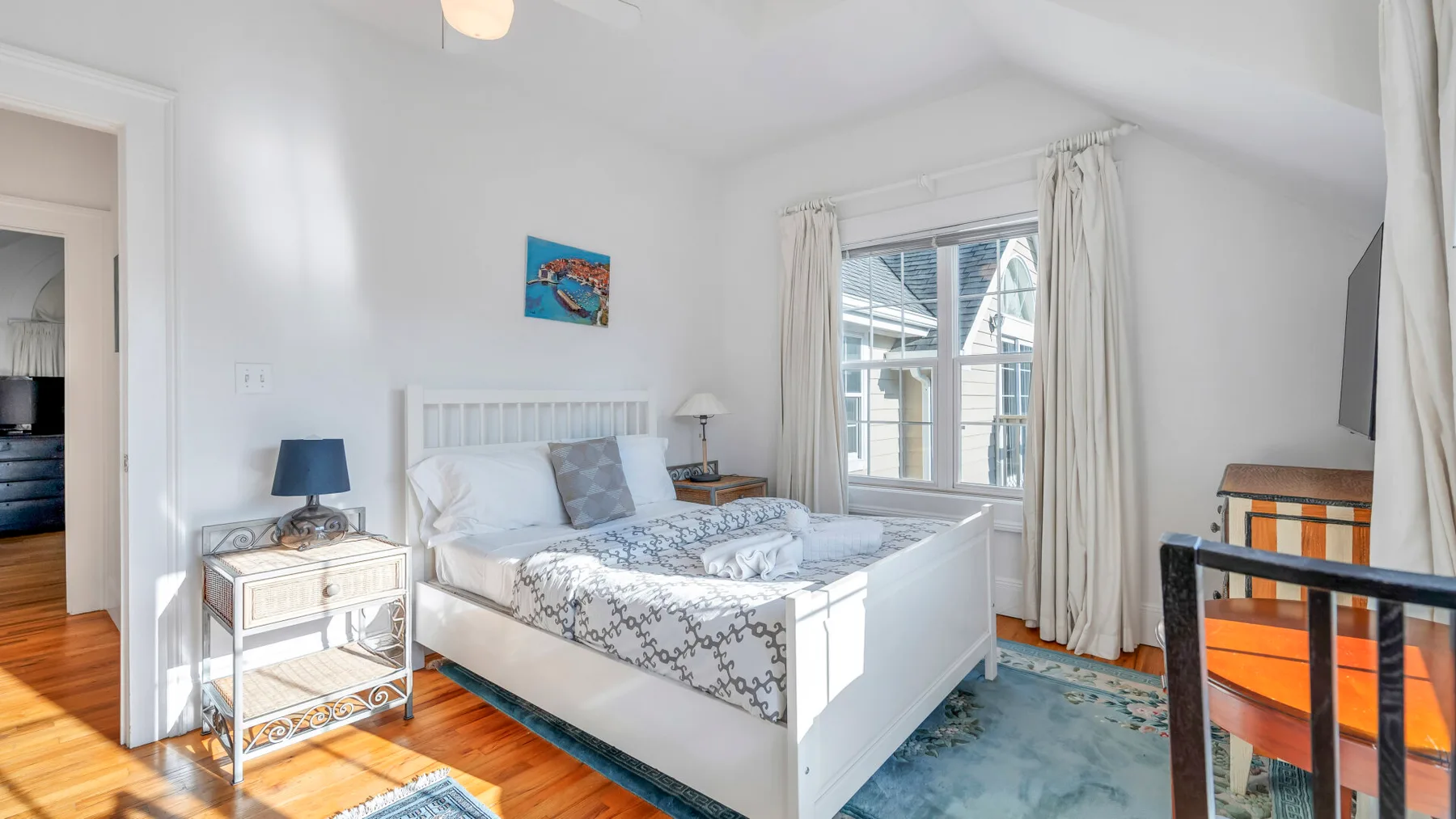 Un dormitorio luminoso con una cama blanca, ropa de cama estampada y una ventana grande.