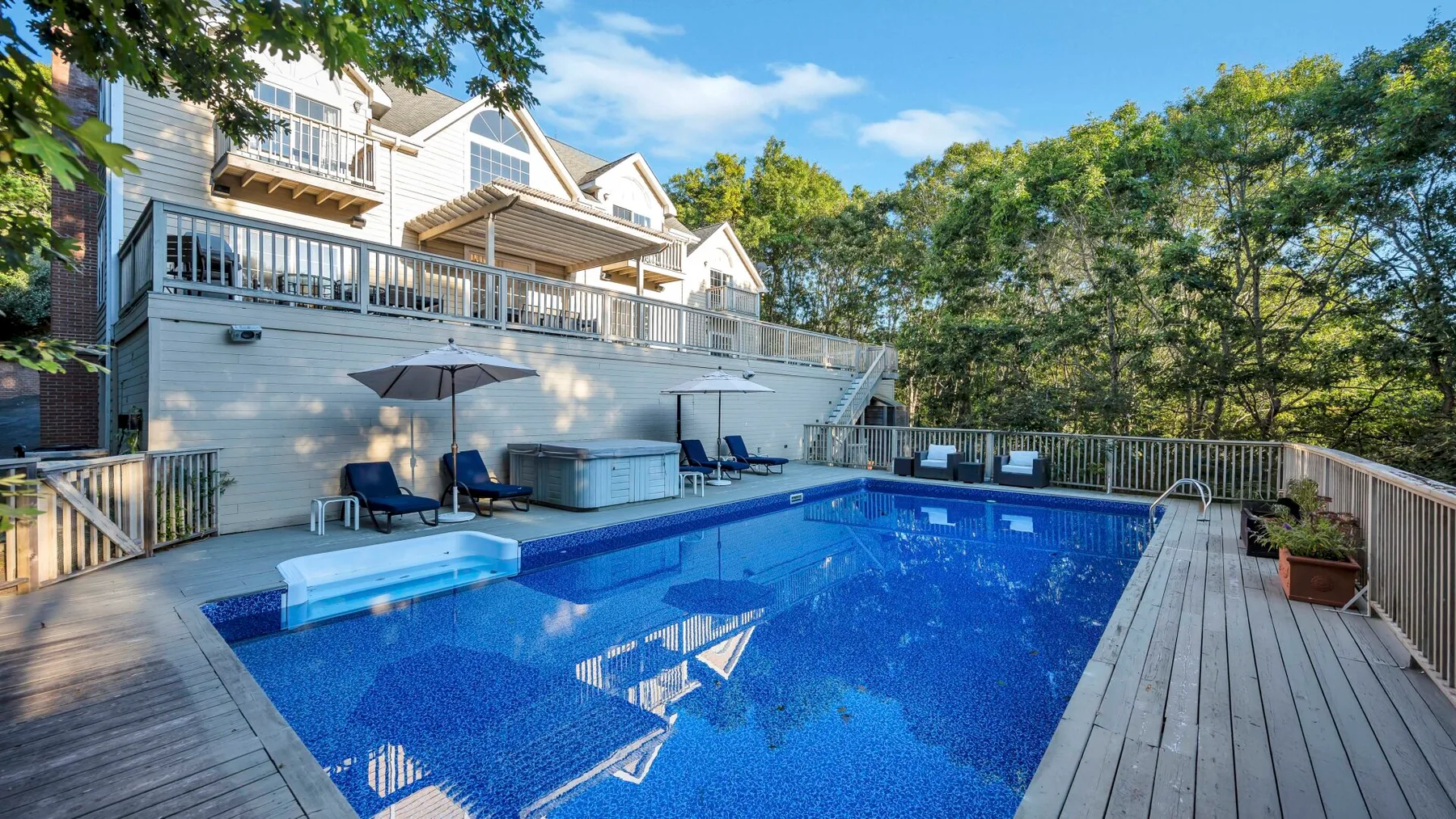 Una piscina al aire libre junto a una terraza, con árboles y una casa grande cerca.