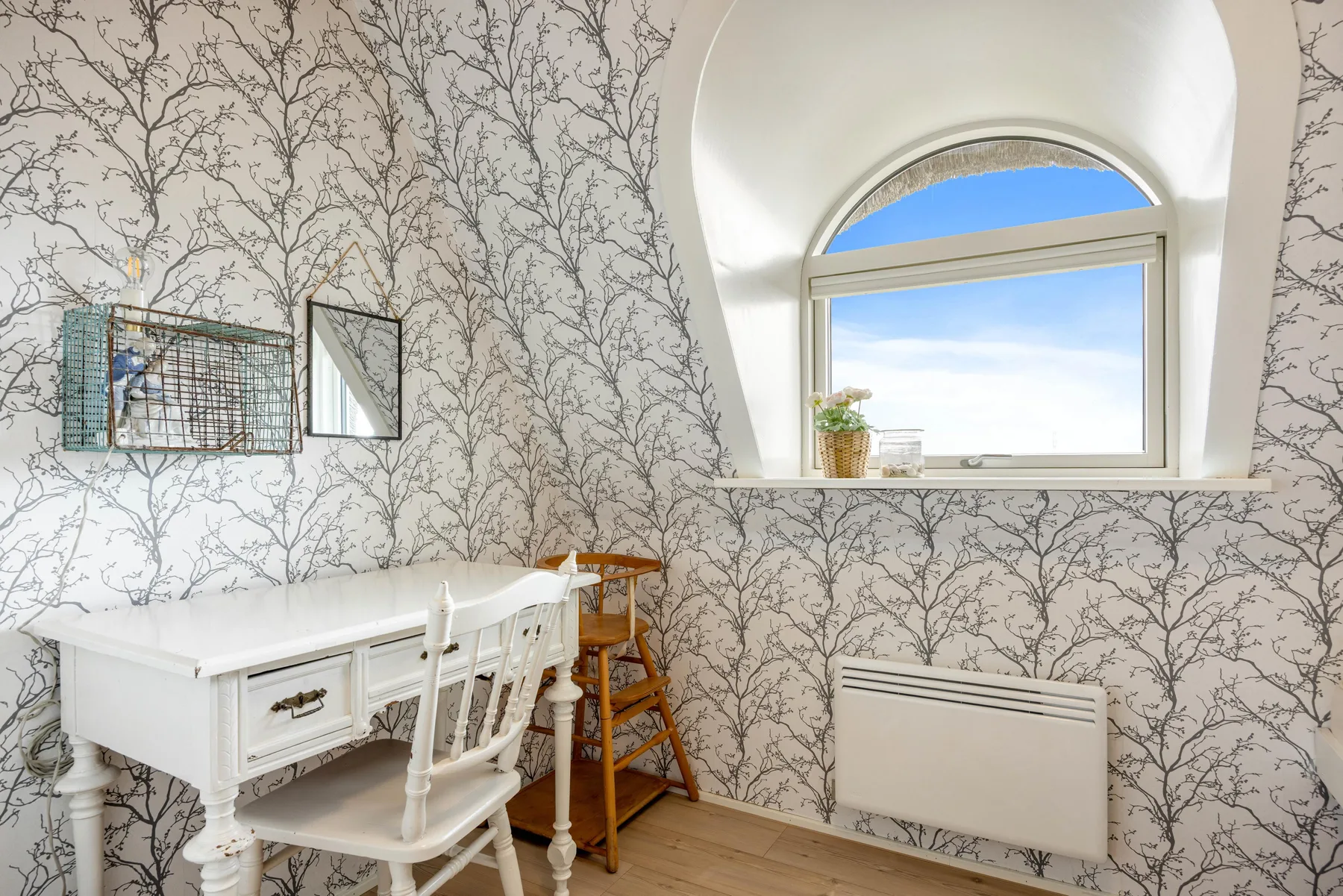 Una habitación luminosa con un escritorio blanco bajo una gran ventana arqueada.