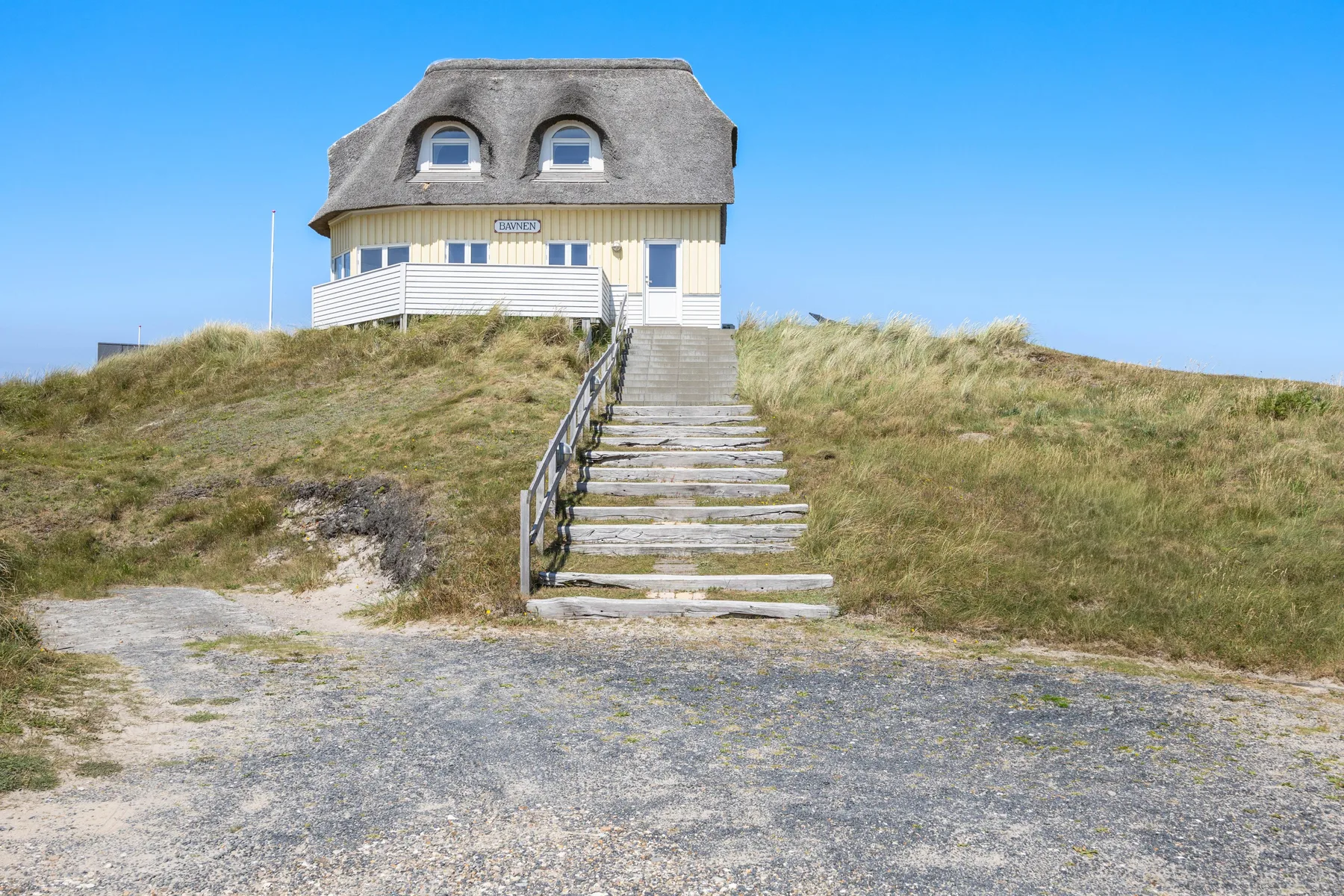 Una casa junto a la playa está sobre dunas cubiertas de hierba, con una escalera de madera.