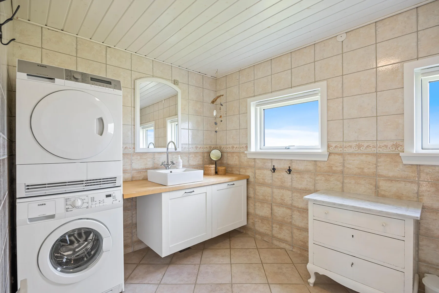 Un baño con azulejos, lavabo, electrodomésticos de lavandería apilados y una cómoda.