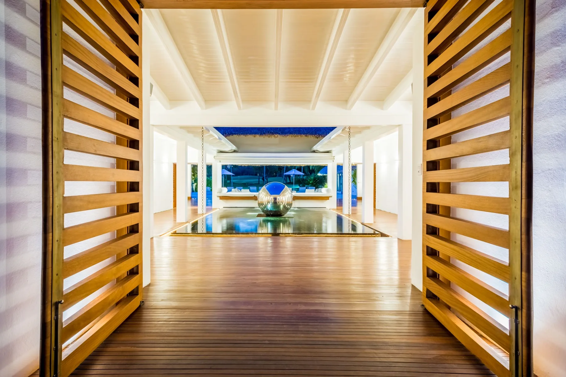Un pasillo de madera conduce entre paneles de listones hacia una piscina reflectante.
