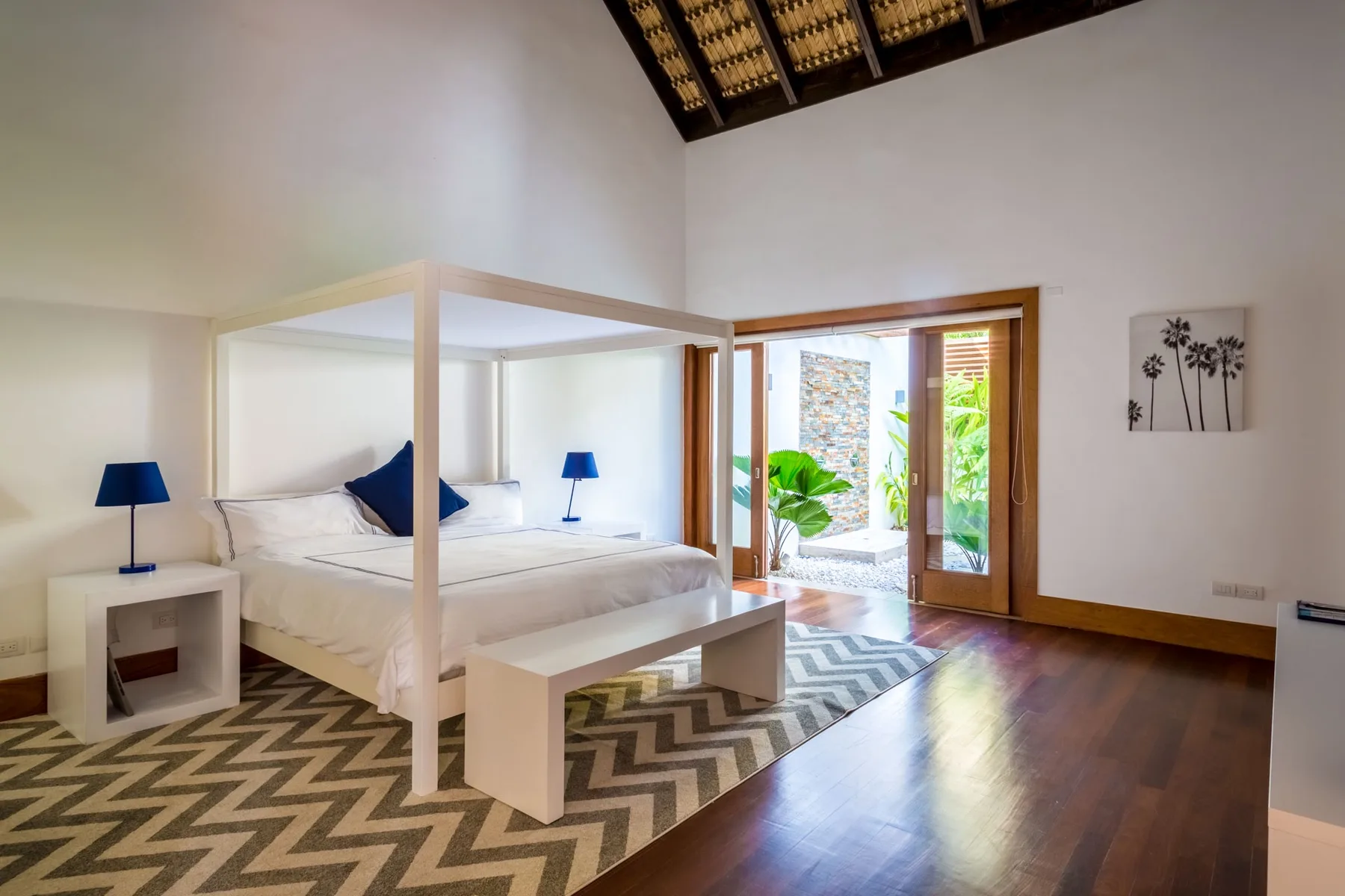 Un dormitorio con cama con dosel, lámparas de noche y puertas de madera abiertas a la vegetación.