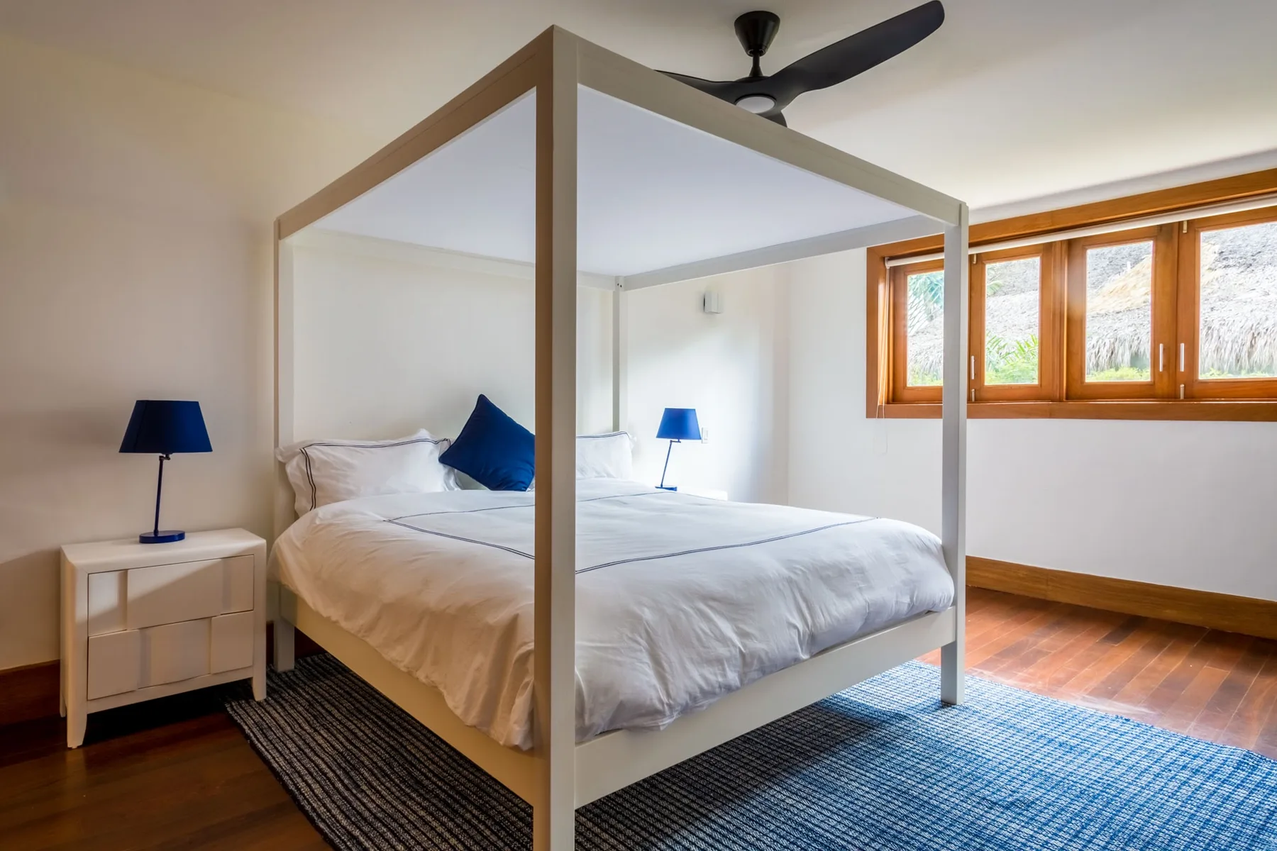 Un dormitorio luminoso con una cama con dosel, mesitas de noche y ventanas en una pared.