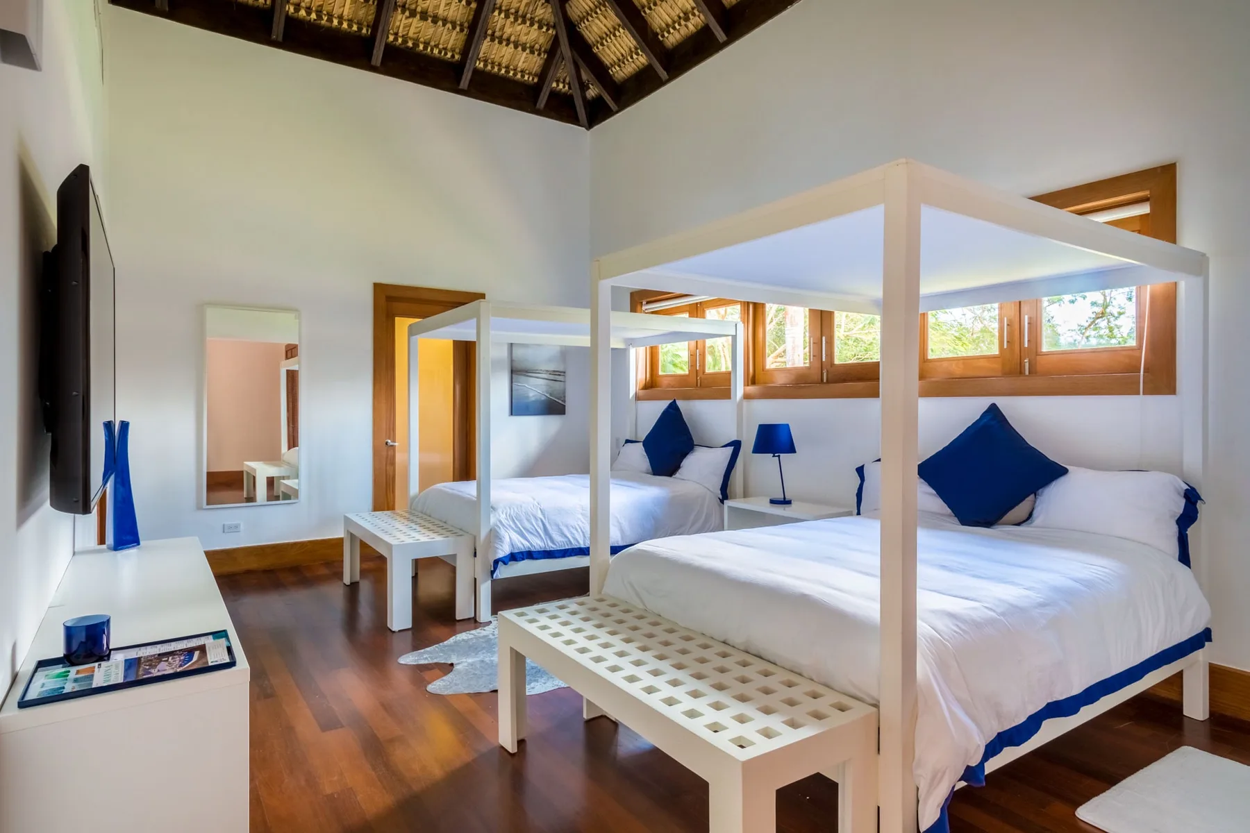Un dormitorio luminoso con camas blancas, detalles azules y ventanas con marcos de madera.