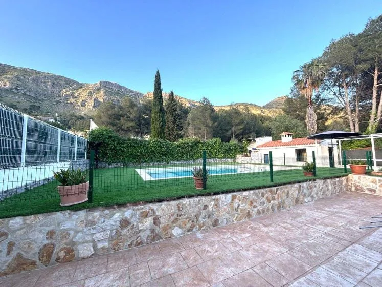 An outdoor swimming pool sits behind a fence, with mountains and trees in the background.