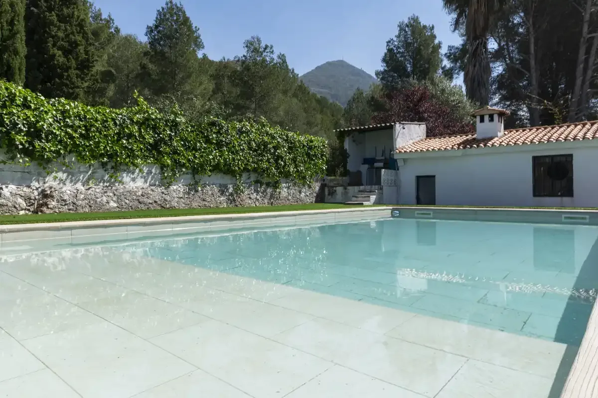 An outdoor swimming pool beside a low white building and leafy trees.