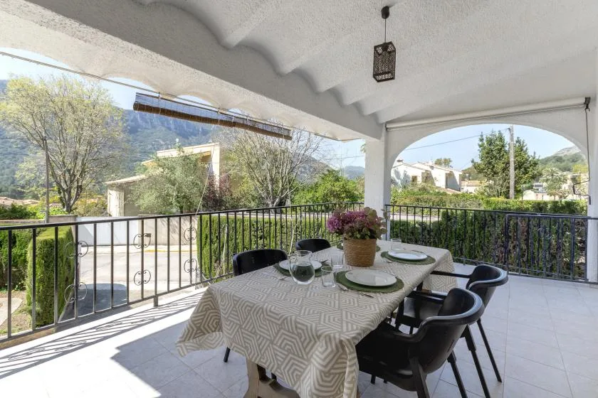 A covered outdoor dining area has a table set beside a railing overlooking greenery.