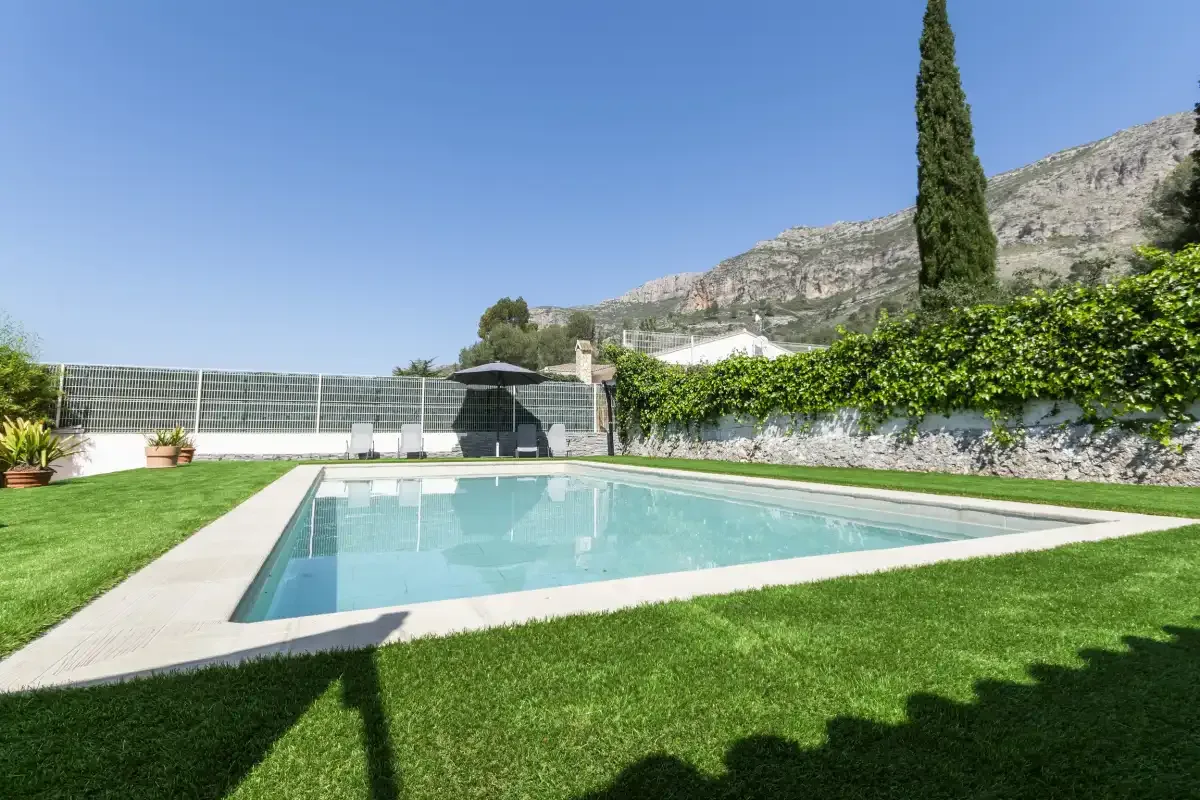 An outdoor swimming pool sits beside a green lawn, with hills in the background.