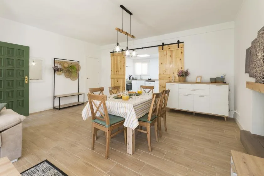 A dining area with a set table, wooden chairs, and a kitchen beyond.