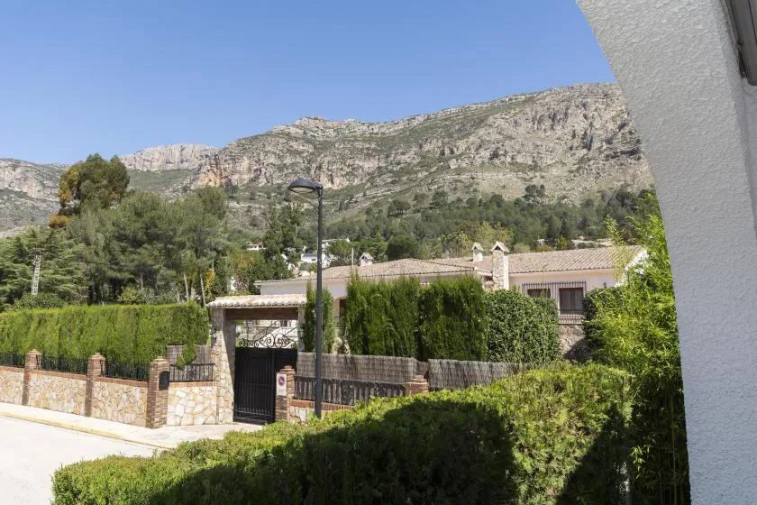A gated residence sits behind hedges, with rocky mountains rising in the background.