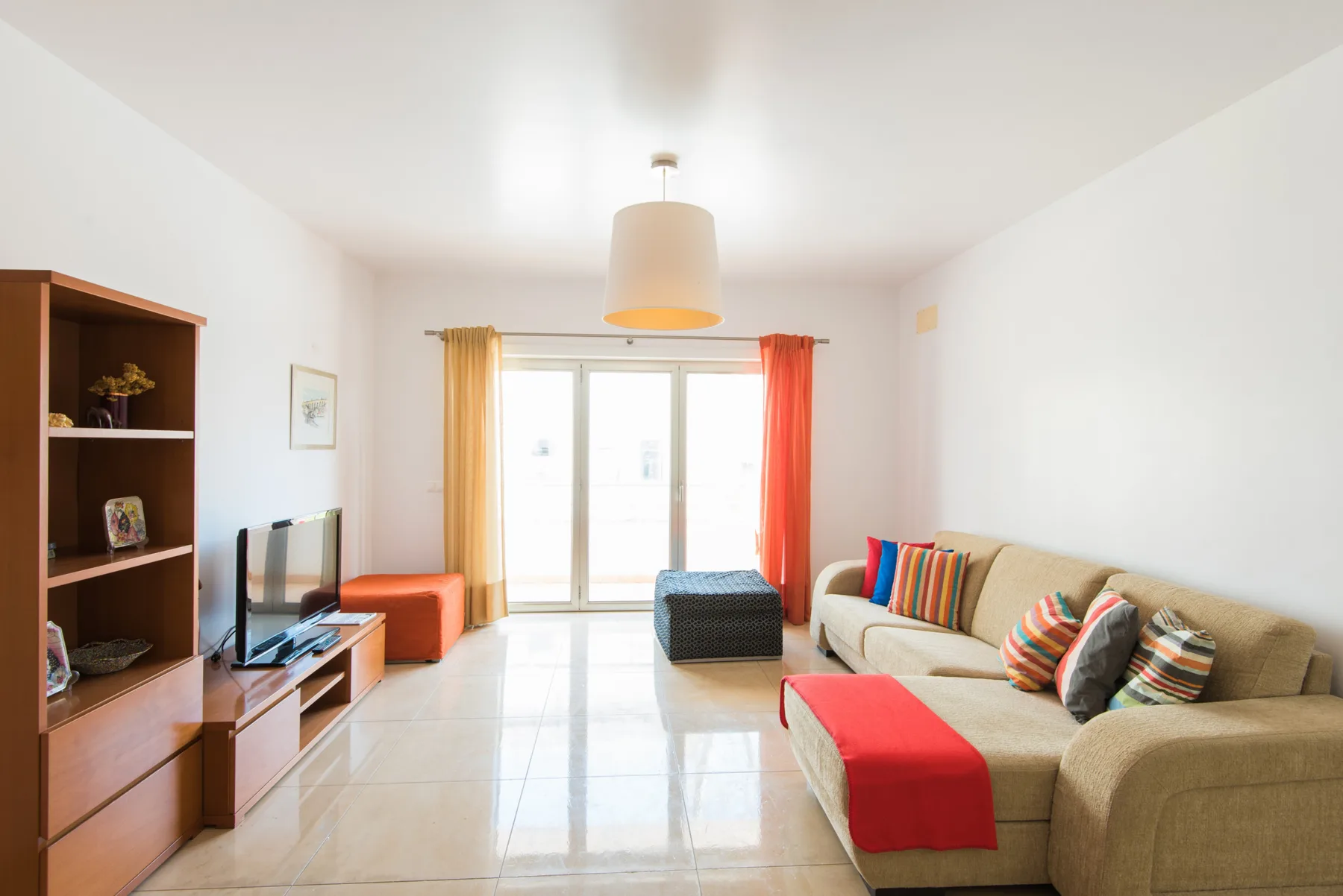 Una sala luminosa con asientos, un televisor y puertas de vidrio que dan a un balcón.