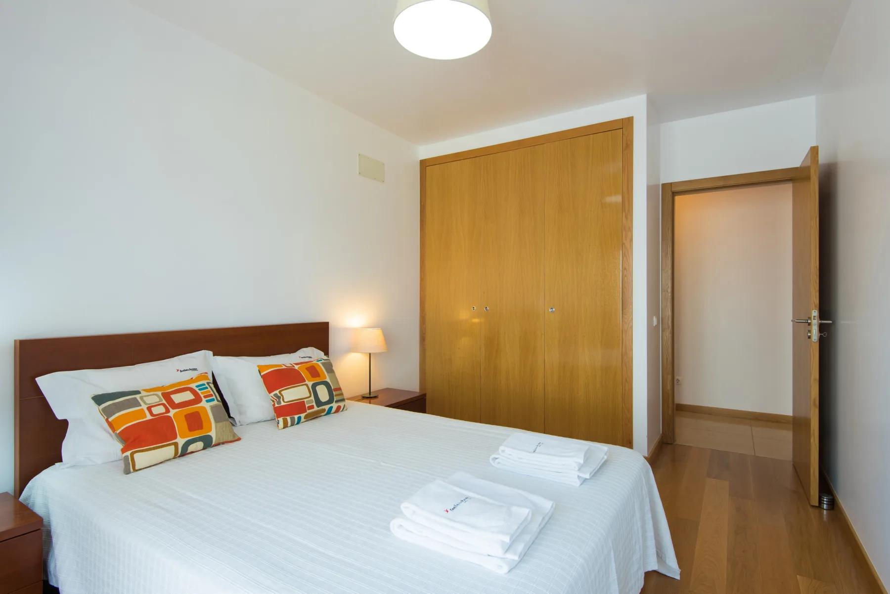 A bedroom with a double bed, folded towels, and wooden wardrobe.