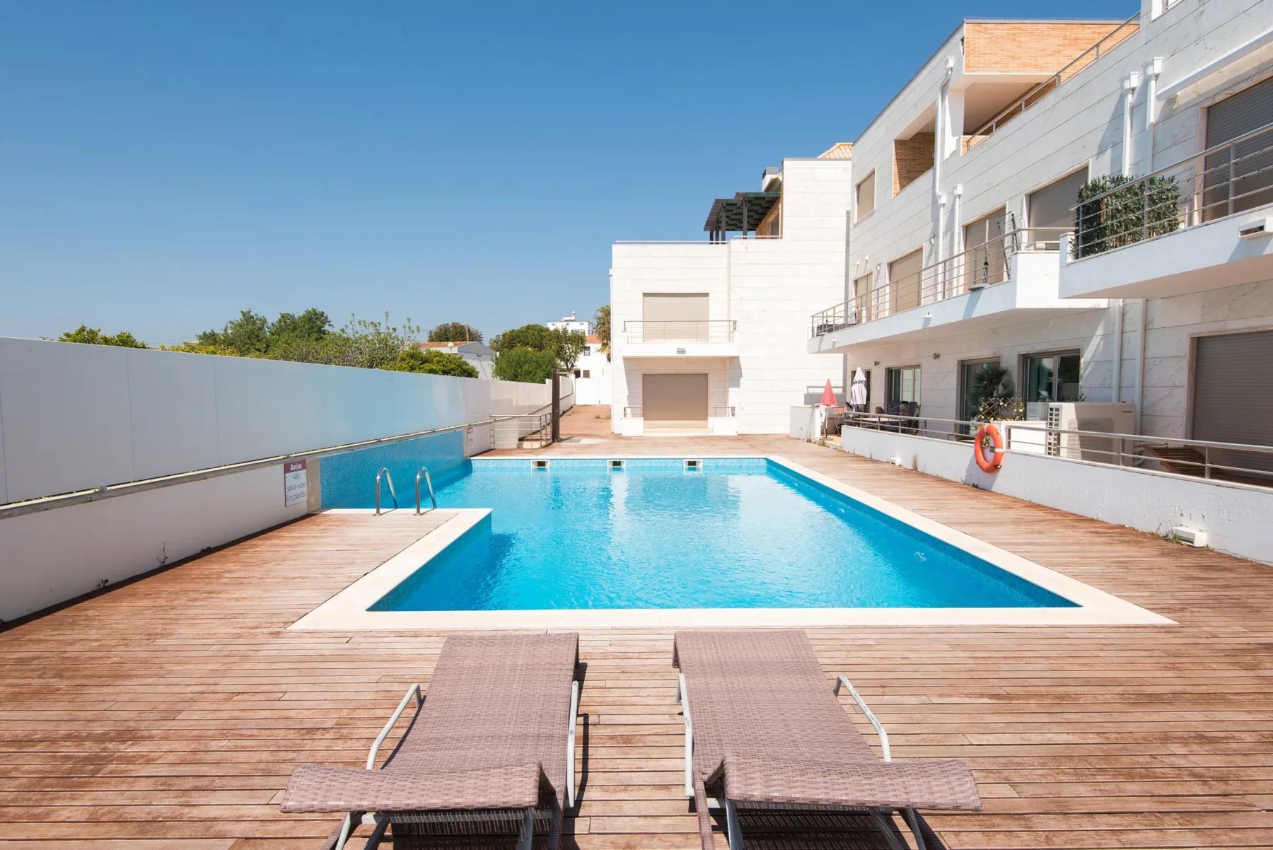 An outdoor swimming pool sits beside a sunlit wooden deck and modern buildings.