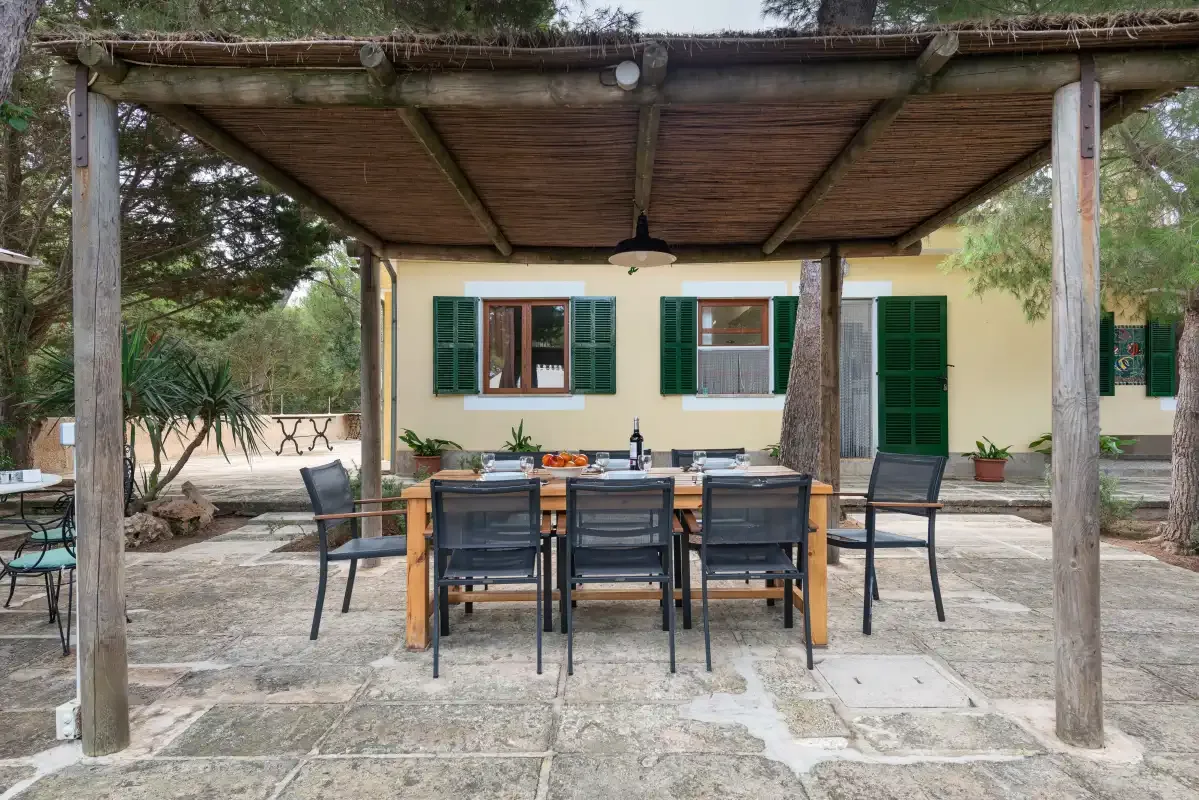 Una zona de comedor exterior está bajo una pérgola de madera junto a una casa.