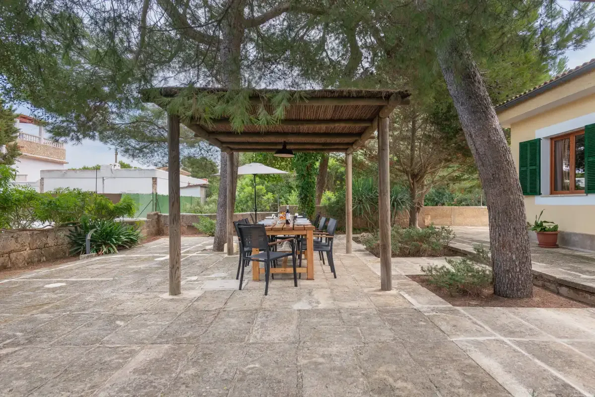 Una zona de comedor exterior pavimentada está bajo una pérgola de madera entre árboles.
