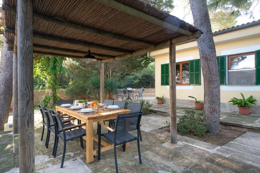 Una zona de comedor al aire libre está bajo una pérgola de madera junto a una casa.