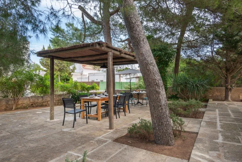 Una zona de comedor al aire libre está bajo una pérgola de madera, entre árboles y arbustos.