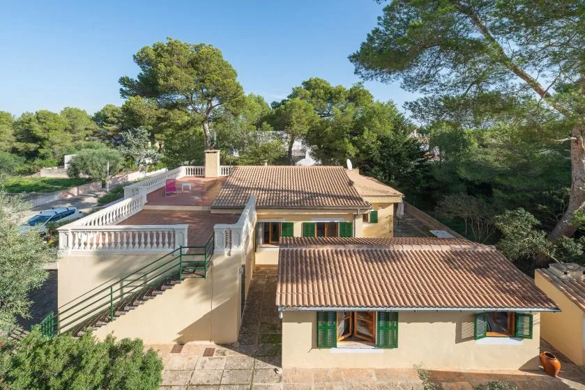 Una casa con techo de tejas y contraventanas verdes está rodeada de árboles altos.