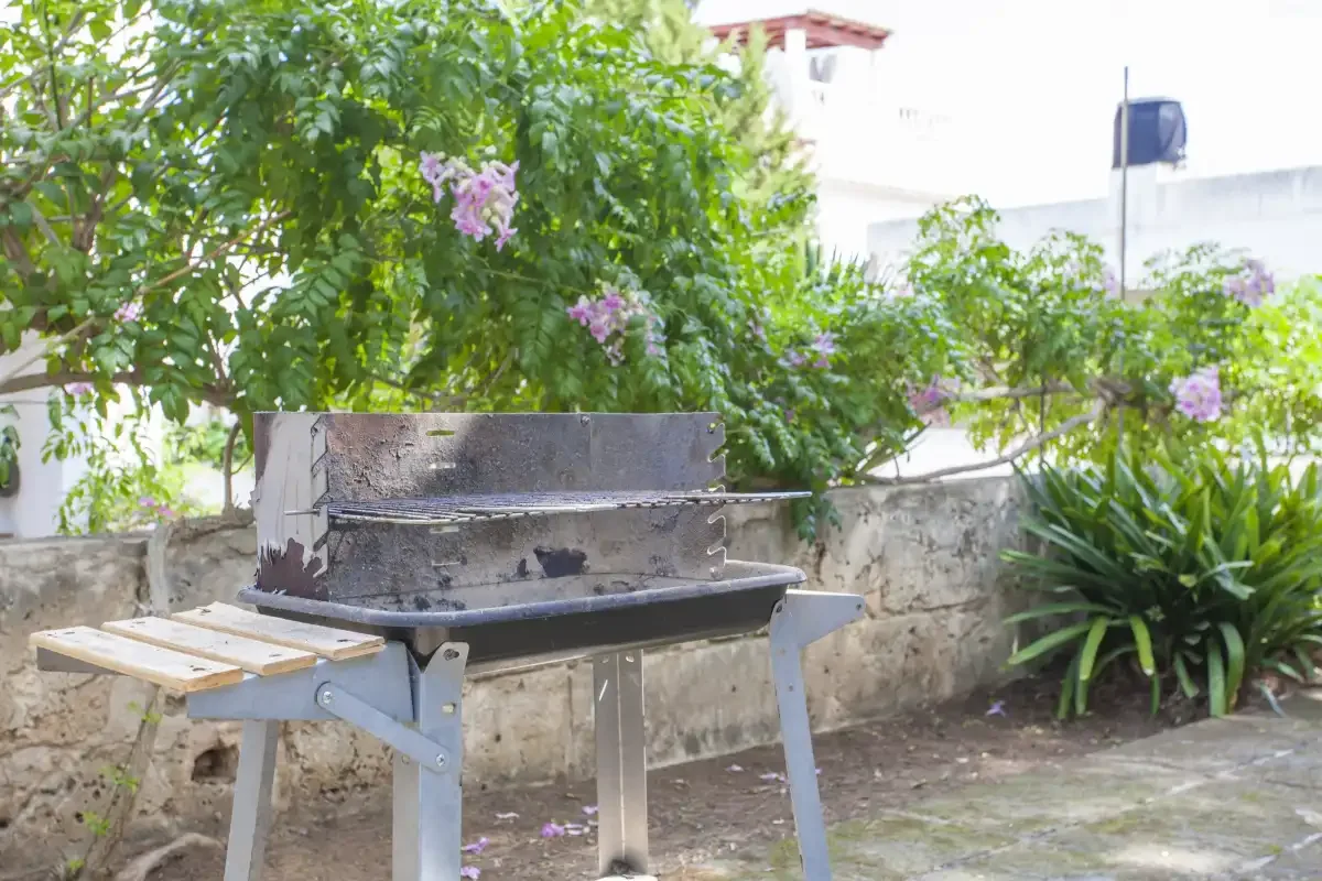 Una parrilla exterior está entre plantas frondosas junto a un muro bajo de piedra.