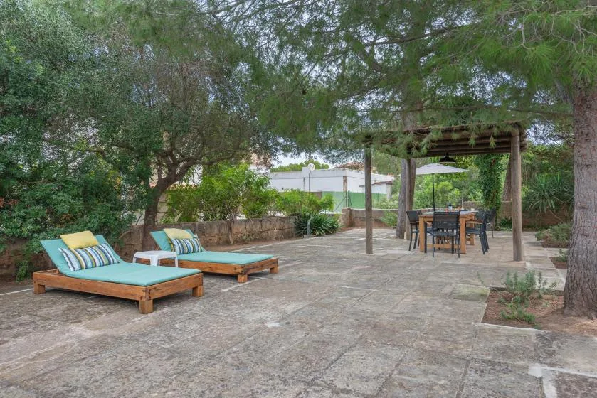 Un patio al aire libre tiene tumbonas acolchadas y una zona de comedor a la sombra de los árboles.