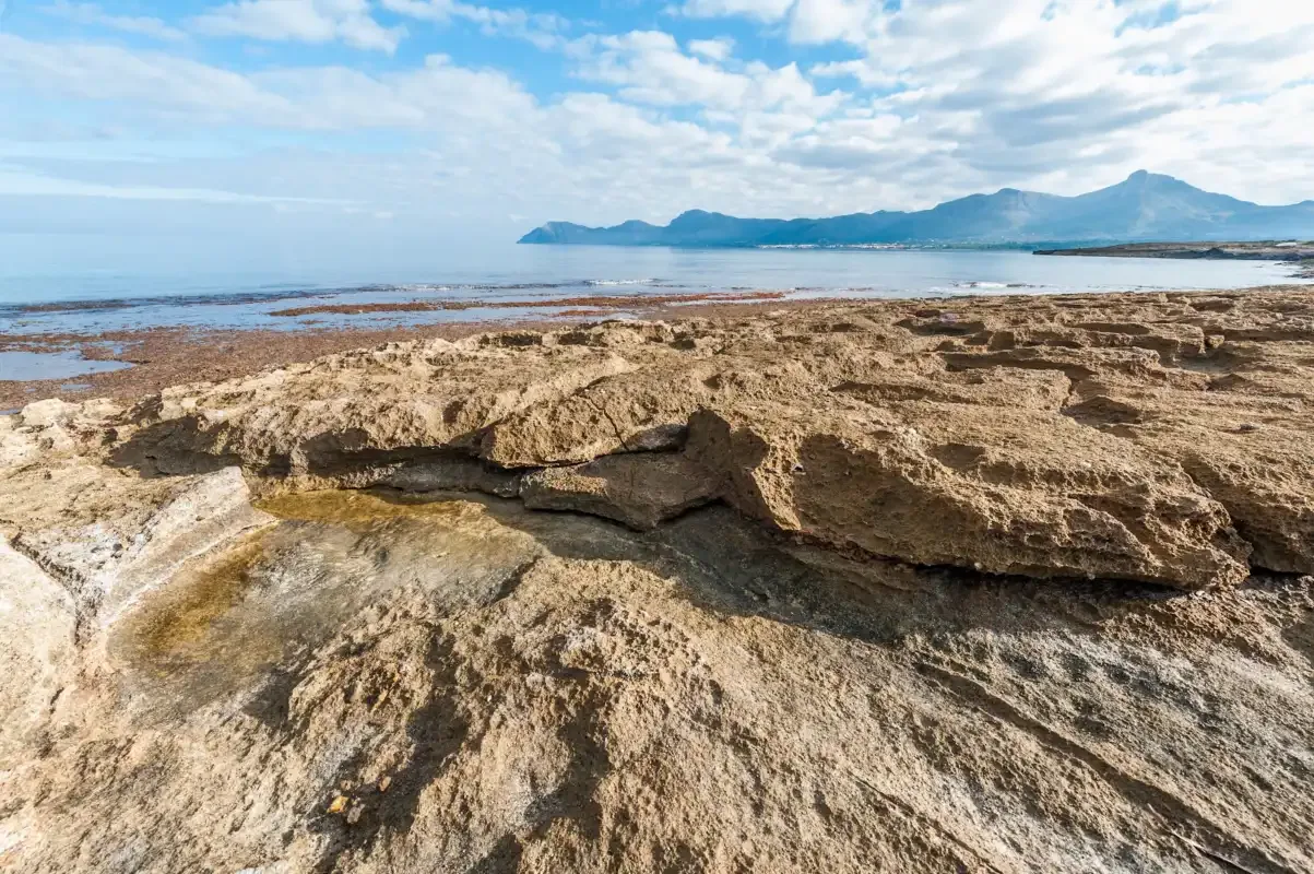 Una costa rocosa bordea aguas poco profundas, con montañas lejanas bajo un cielo parcialmente nublado.