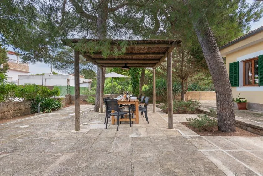 Una zona de comedor exterior pavimentada se encuentra bajo una pérgola de madera entre árboles.