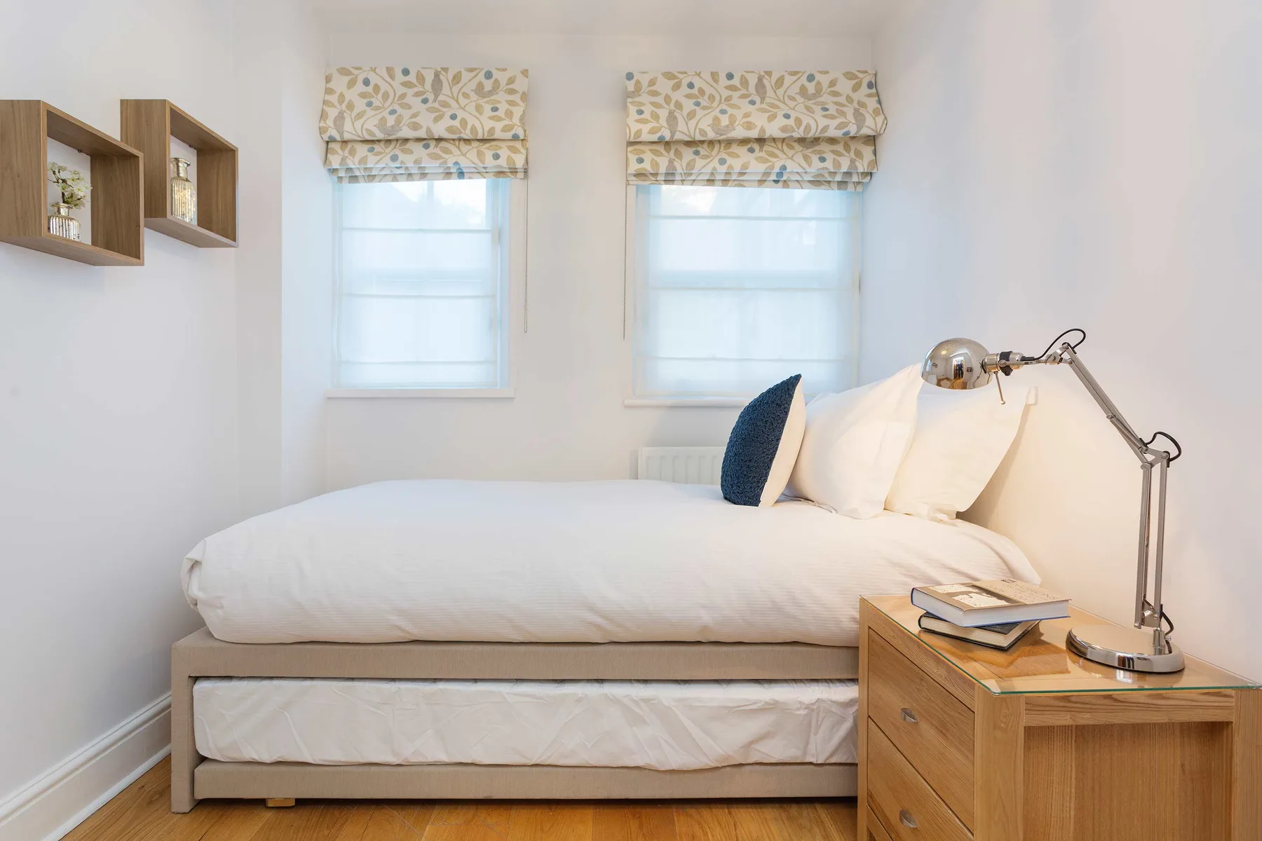 A bright bedroom with a bed beneath two windows and a bedside table.