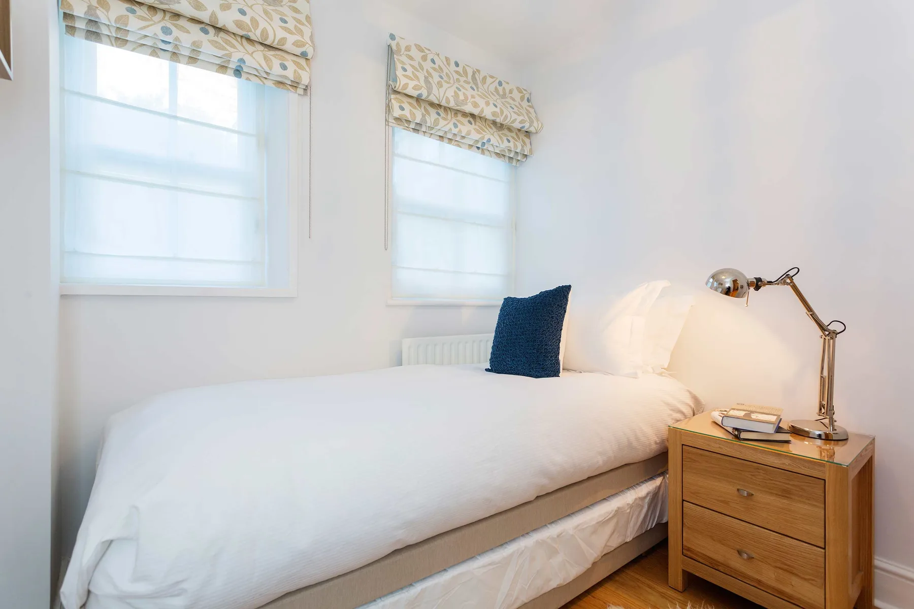 A bright bedroom with a white bed, patterned window shades, and a bedside table.