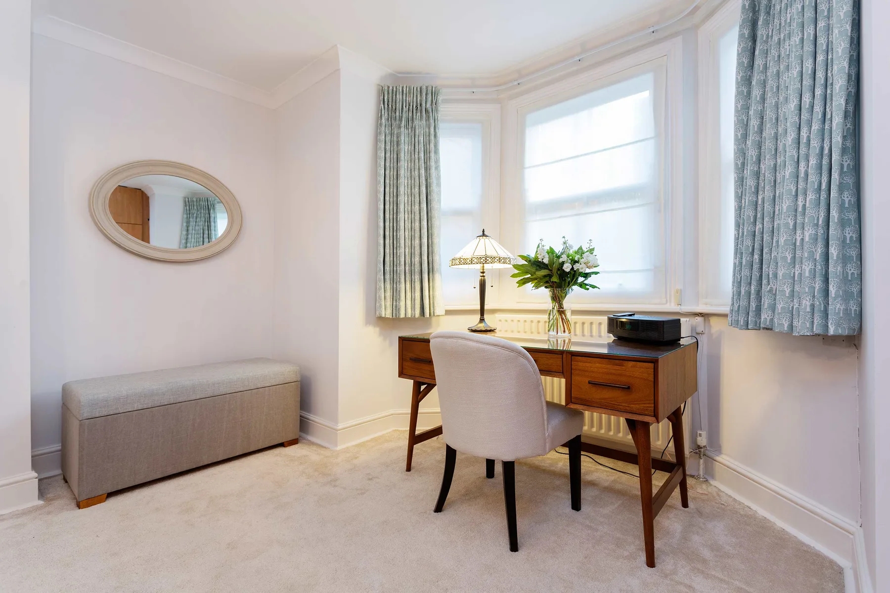 A bright room features a wooden desk beneath bay windows and a cushioned seat.
